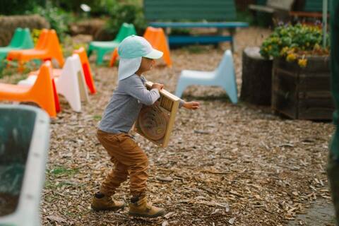 A toddler in a bucket hat carrying a board past the kids' chairs